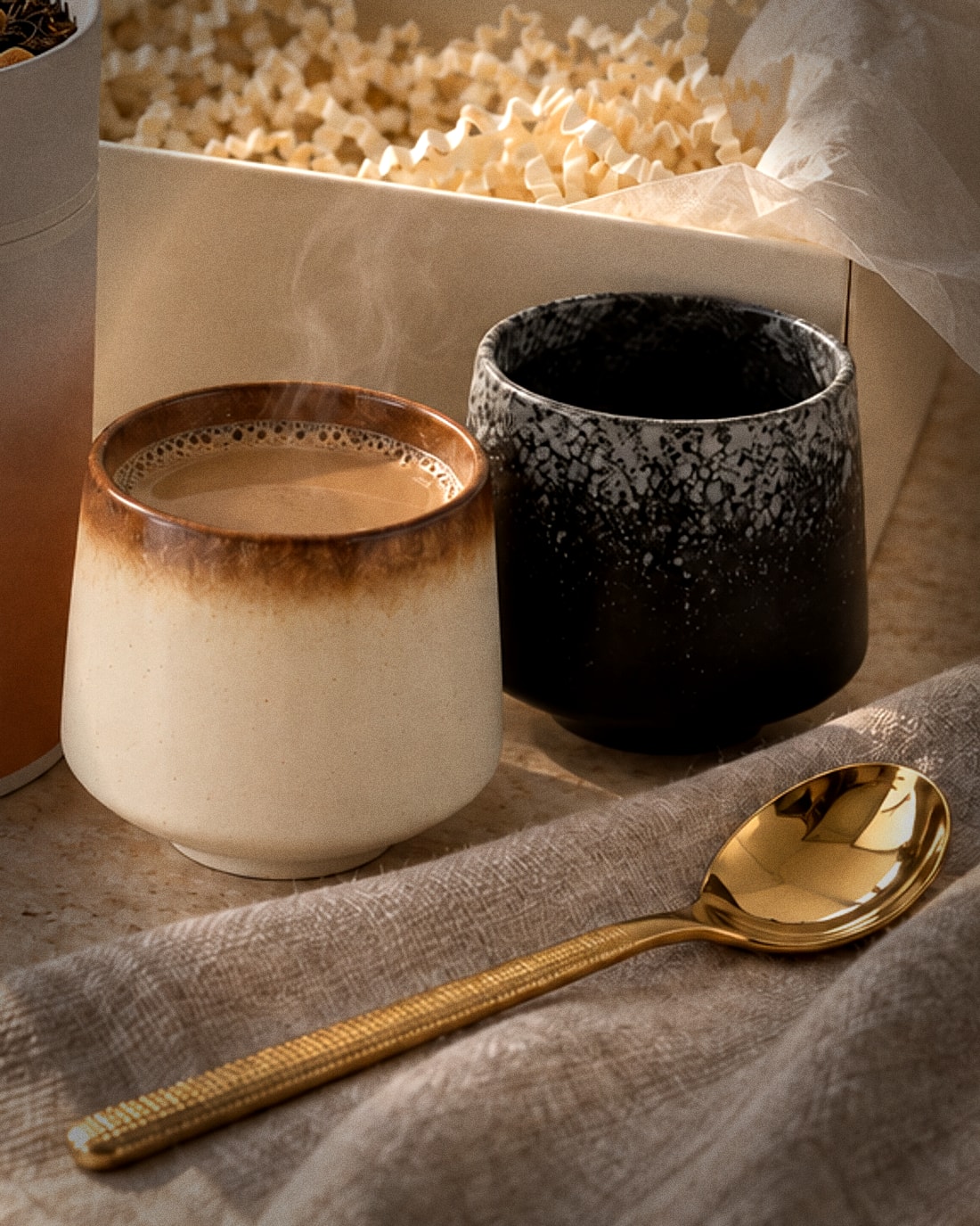 A cream ceramic cup of steaming chai beside a speckled black cup, with a golden spoon on linen